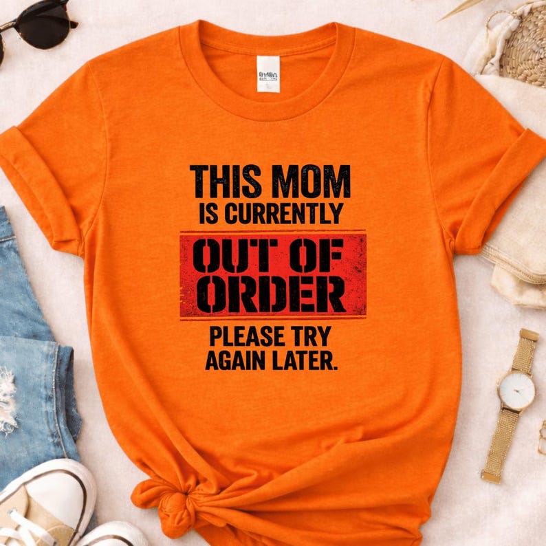 Pode incluir: Camiseta laranja com o texto "THIS MOM IS CURRENTLY OUT OF ORDER PLEASE TRY AGAIN LATER" em preto e vermelho. A camiseta tem mangas curtas e gola redonda.