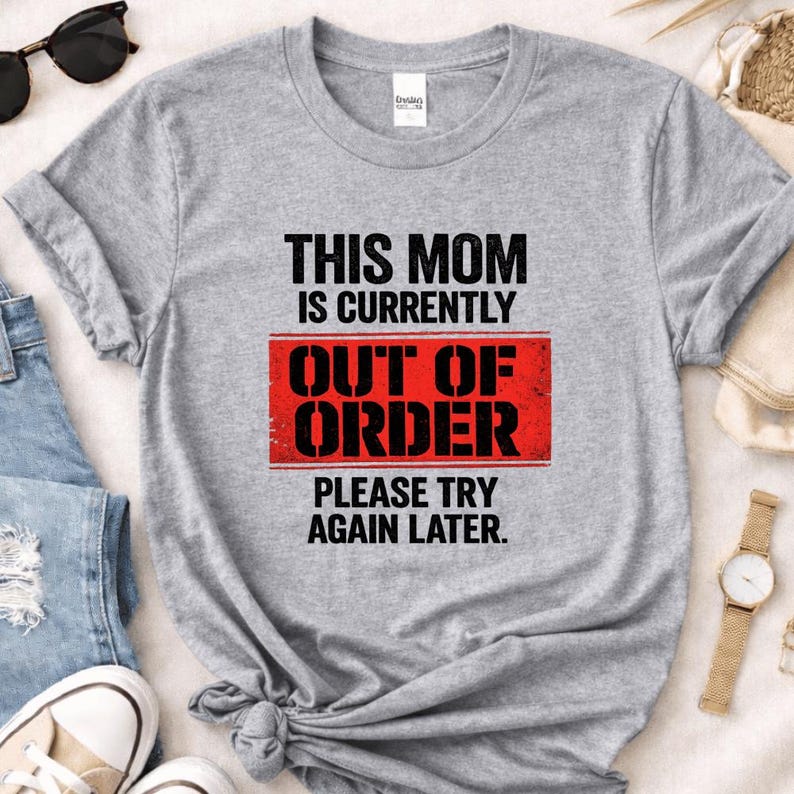 Pode incluir: Uma camiseta cinza mesclada com o texto "THIS MOM IS CURRENTLY OUT OF ORDER PLEASE TRY AGAIN LATER" em preto e vermelho. A camiseta tem gola redonda e parece ser feita de um material macio. &Oacute;culos de sol e um rel&oacute;gio s&atilde;o vis&iacute;veis no fundo.