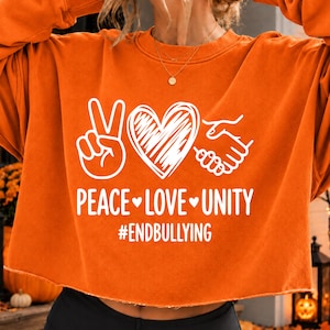 May include: An orange long-sleeve sweatshirt featuring a white graphic of a peace sign, a heart, and a handshake. Below the graphic, the text reads PEACE LOVE UNITY and #ENDBULLYING in white capital letters. The sweatshirt has a crew neckline.