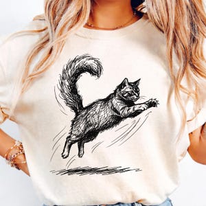 May include: A cream-colored t-shirt featuring a black ink-style sketch of a fluffy cat mid-leap. The cat is depicted with a long, arched, bushy tail and extended front paws, captured in a dynamic motion with sketchy lines underneath its feet.