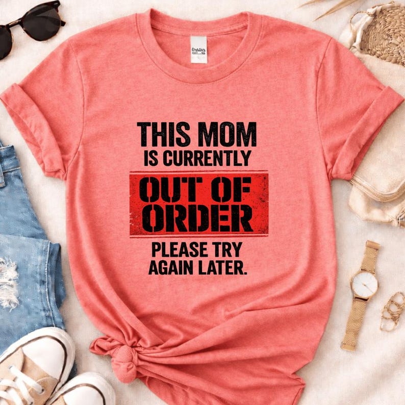 Pode incluir: Uma t-shirt cor-de-coral com o texto "THIS MOM IS CURRENTLY OUT OF ORDER PLEASE TRY AGAIN LATER" em preto e vermelho. A t-shirt tem mangas enroladas e um n&oacute; na parte inferior. &Oacute;culos de sol, um rel&oacute;gio e t&eacute;nis s&atilde;o vis&iacute;veis.