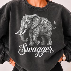 May include: A charcoal gray crewneck sweatshirt featuring a white graphic of an elephant decorated with intricate floral and scroll patterns. Below the elephant, the word "Swagger" is printed in a white, cursive, decorative script font.