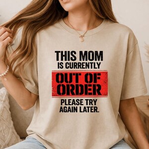 Pode incluir: Camiseta bege com o texto "THIS MOM IS CURRENTLY OUT OF ORDER PLEASE TRY AGAIN LATER" em preto e vermelho. A camiseta tem gola redonda e mangas curtas.