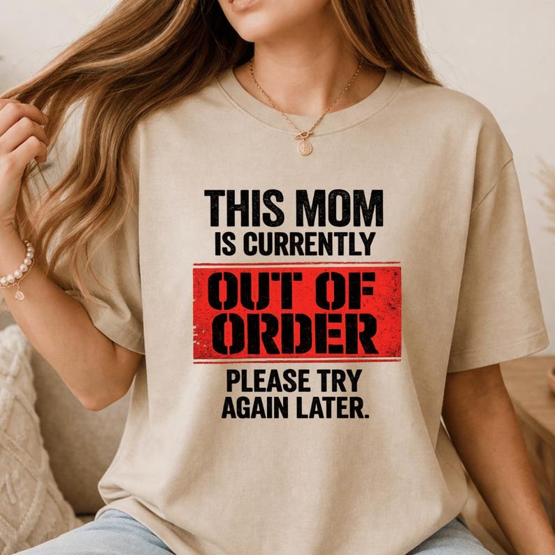 Pode incluir: Camiseta bege com o texto "THIS MOM IS CURRENTLY OUT OF ORDER PLEASE TRY AGAIN LATER" em preto e vermelho. A camiseta tem gola redonda e mangas curtas.
