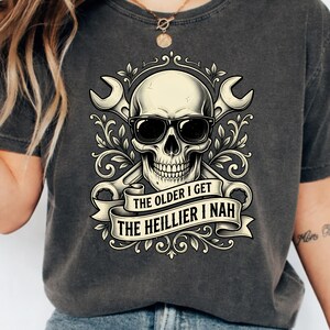 Op de afbeelding: Een antracietgrijs t-shirt met een afbeelding van een schedel met een zonnebril voor twee gekruiste moersleutels. Onder de schedel staat een banner met de tekst THE OLDER I GET THE HEILLIER I NAH, omringd door decoratieve krulmotieven.