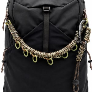 May include: A black backpack featuring a decorative paracord attachment in shades of olive green, brown, and tan. The cord is woven with several small, light green circular rings and includes a black metal carabiner and a dangling cord pull.