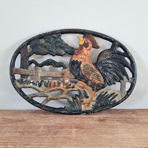 May include: An oval cast iron wall plaque featuring a relief design of a rooster standing on a wooden fence. The rooster is painted in shades of brown, orange, and black. The background includes a tree and mountain landscape in muted green and black tones.