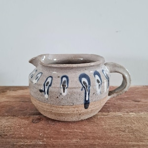 May include: A handmade ceramic creamer with a spout and handle. The stoneware creamer is light grey with dripped blue and white accents. The base is a lighter tan colour. The creamer sits on a wooden surface.