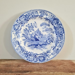 May include: A round ceramic plate featuring a blue and white transferware pattern. The centre depicts a landscape with a stone building and trees, while the wide border is decorated with intricate floral motifs and foliage.