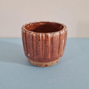 May include: A small, cylindrical ceramic cup featuring a textured, reddish-brown speckled glaze. The exterior surface is defined by vertical fluted grooves running along the sides, while the base remains an unglazed, light tan clay finish.