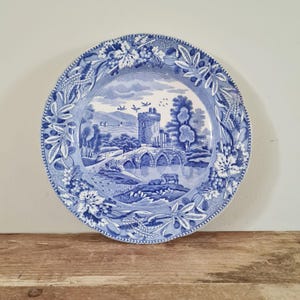 May include: A round ceramic plate featuring a blue and white transferware pattern. The centre depicts a landscape with a stone castle, a multi-arched bridge over water, and grazing cattle. The border is decorated with grapevines and wheat stalks.