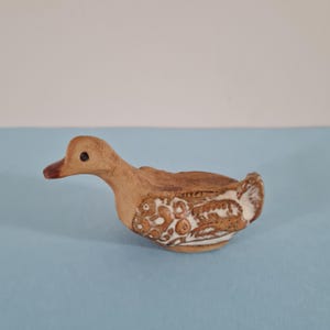 May include: A small, ceramic duck figurine in shades of brown and white. The duck has a detailed design on its body, with a light brown head and a darker brown beak. The figurine is a decorative item.