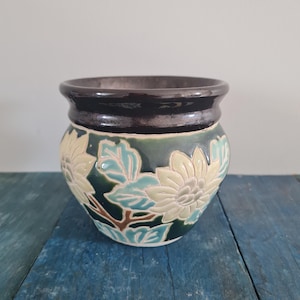 May include: A ceramic flower pot with a dark brown rim and a green base. The pot is decorated with cream-colored floral designs and light blue leaves. The pot sits on a blue wooden surface.