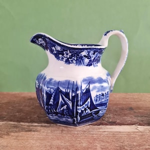 May include: A white ceramic cream jug featuring a blue transferware pattern. The body displays a detailed nautical scene with sailboats and figures, while the rim is decorated with a floral border. It has a curved handle and a pouring spout.