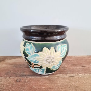 May include: A ceramic planter featuring a dark brown rim and a deep green body decorated with embossed, light-colored floral patterns and leaves. The pot has a rounded shape with a wide opening and a smooth, glossy finish throughout the exterior.