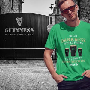 May include: A green t-shirt with the text "Hello Darkness My Old Friend, I've Come to Drink With You Again" and three dark beer glasses with shamrocks. The background features the Guinness brewery gates.