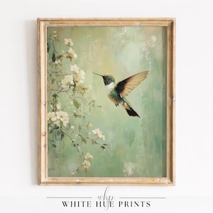 Puede incluir: Una impresión enmarcada que presenta un colibrí en vuelo, con las alas extendidas, sobre un fondo suave verde y blanco. Flores blancas florecen en una rama en primer plano. El marco es de color dorado claro y desgastado. El texto en la parte inferior dice "WHITE HUE PRINTS."