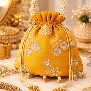 Może przedstawiać: Musztardowa torba ze sznurkiem ze haftem kwiatowym i perłowymi akcentami. Torba posiada uchwyt z koralików i zwisające frędzle. Inna złota i perłowa biżuteria znajduje się w tle.