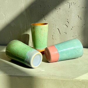 Puede incluir: Tres vasos de cerámica en tonos verdes, azules y rosas. Los vasos tienen una textura moteada y un acabado brillante. Un vaso está en posición vertical, mientras que los otros dos están tumbados de lado. El interior de un vaso es naranja.