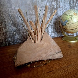 Könnte beinhalten: Eine handgefertigte Holzvase mit dreieckiger Form, die mehrere Holzstäbe und grüne Zweige enthält. Die Vase steht auf einem goldfarbenen Sockel. Im Hintergrund ist ein Globus zu sehen.