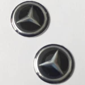 May include: Two circular emblems featuring a silver three-pointed star logo centered on a black background, surrounded by a polished silver-toned metallic ring. These round automotive-style badges have a smooth, reflective surface finish.