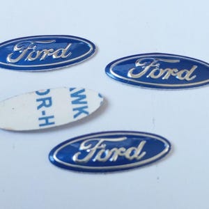 Peut inclure: Trois emblèmes Ford de forme ovale avec un fond bleu brillant et des lettres argentées en relief. Deux emblèmes affichent clairement le logo Ford, tandis qu'un emblème est retourné pour révéler un support adhésif blanc avec le texte bleu WK OR-H.