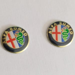 Może przedstawiać: Dwa okrągłe emblematy Alfa Romeo z czarnym zewnętrznym pierścieniem i złotymi literami. Środek przedstawia czerwony krzyż na białym tle oraz zielonego węża na niebieskim tle, całość zamknięta w cienkiej, złotej metalowej obwódce.