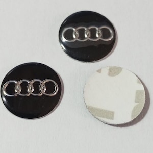 Könnte beinhalten: Drei kreisförmige Embleme mit dem silbernen Audi-Vier-Ringe-Logo auf glänzend schwarzem Hintergrund. Zwei Embleme sind von vorne zu sehen, während das dritte Emblem umgedreht ist und seine weiße Kleberückseite zeigt.