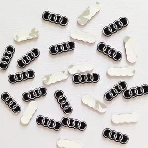 Peut inclure: Une collection de petits emblèmes individuels avec le logo Audi, composés de quatre anneaux entrelacés noirs et argentés. Certains emblèmes sont présentés avec le logo noir et argenté vers le haut, tandis que d'autres montrent le dos argenté réfléchissant.