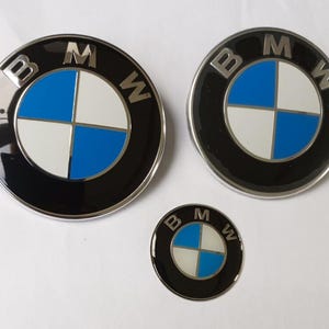 Op de afbeelding: Drie BMW-emblemen in verschillende maten. Elk embleem heeft een zwarte buitenring met de letters "BMW" en een centraal cirkelvormig ontwerp verdeeld in vier blauwe en witte kwadranten.