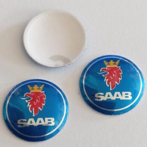 May include: Three circular emblems featuring the Saab logo. Two emblems are blue with a red griffin head wearing a gold crown and the white text SAAB. One emblem is plain white on the front side, showing a smooth, rounded, and glossy surface.