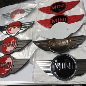 Könnte beinhalten: Eine Sammlung von MINI-Autoemblemen in verschiedenen Farben, darunter Rot, Schwarz, Silber und Chrom. Jedes Emblem zeigt das Wort "MINI" in einem kreisförmigen Design mit Flügelakzenten. Die Embleme sind neu und verpackt.