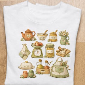 May include: White t-shirt featuring a watercolor illustration of kitchen items. The design includes a teapot, honey jar, scale with apples, pasta jar, butter dish, oil bottles, and an apron. The design is in soft, muted colors.