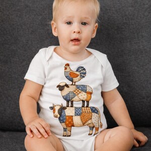 Puede incluir: Body de bebé blanco con un diseño caprichoso de un pollo, una oveja y una vaca apilados. Los animales tienen un patrón de patchwork en tonos azules, marrones y naranjas. El body es de manga corta y tiene cuello redondo.