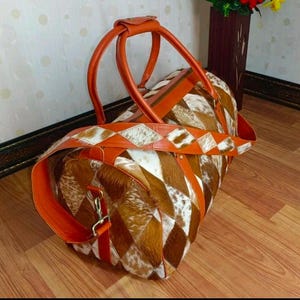 May include: A travel duffel bag with a patchwork design of brown, white, and tan cowhide. It has orange leather handles, a shoulder strap, and diamond-shaped accents. The bag is designed for travel.