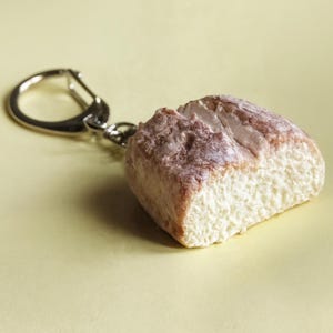 Pode incluir: Um porta-chaves em forma de pão. O porta-chaves apresenta uma pequena e detalhada réplica de um pão com um exterior crocante e texturizado e um interior claro e poroso. O porta-chaves está preso a um anel de metal prateado.