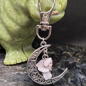 May include: Silver-toned keychain featuring a crescent moon pendant with intricate filigree detailing. The moon holds several small, pink rose quartz stones. The keychain has a lobster clasp for easy attachment.