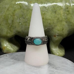 May include: A silver ring with a turquoise oval stone, set in a feather design. The ring is displayed on a white cone-shaped stand, with a green sculpture in the background. The ring is a piece of jewelry.
