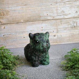 May include: A small, green and black bear figurine with a textured surface. The bear is sitting on a grey surface with green plants on either side. The text "Sunset Meadow Stone Co." is at the bottom.