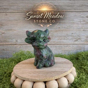 May include: A green and pink stone figurine of a small, seated creature with horns and large ears. The figurine is on a wooden stand with a beaded base, set against a backdrop of wood and greenery. The Sunset Meadow Stone Co. logo is visible.
