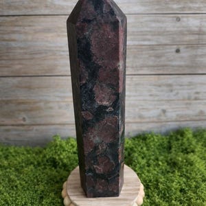 Może przedstawiać: Wysoki, ciemnoczerwony i czarny kamienny obelisk ze spiczastym wierzchołkiem. Kamień osadzony jest na okrągłej drewnianej podstawie z małymi drewnianymi koralikami na krawędzi. Obelisk eksponowany jest na zielonym mchu.