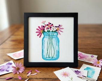May include: A square black frame containing a white fabric panel with an embroidered light blue mason jar filled with pink and purple flowers. The embroidery features green stems and detailed petals, presented in a clean, centered composition.
