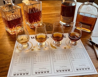 May include: Two clear glass decanters with geometric patterns, two glass bottles of amber liquid, four tulip-shaped tasting glasses filled with spirits, and a white paper tasting mat with printed fields for notes and star ratings for whiskey.