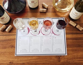 May include: A wine tasting set featuring a rectangular paper mat with four numbered sections for rating wine, four clear glass stems filled with different colored wines, two glass decanters, two bottles of Pinot Noir, one bottle of Chardonnay, and corks.