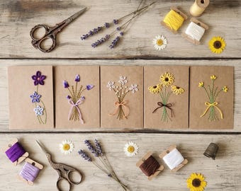 May include: Five brown paper cards featuring hand-embroidered flower bouquets, accompanied by vintage-style metal scissors, spools of colorful embroidery thread, dried lavender sprigs, small white daisy blossoms, and a metal thimble.