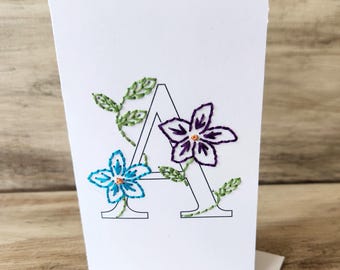 May include: A white card with the letter A embroidered in black thread. The letter is surrounded by green leaves and purple and blue flowers.