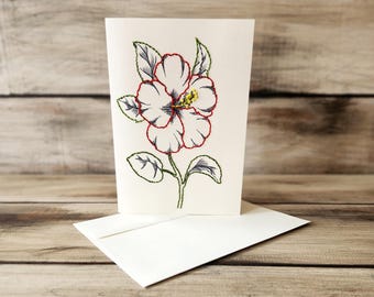 May include: A white card with a hand-stitched hibiscus flower in red, blue, and green thread. The card is blank and comes with a white envelope.