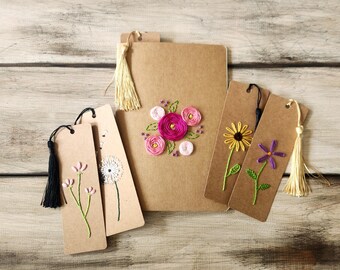 May include: A collection of handmade floral-themed bookmarks and a journal. The bookmarks are made of tan cardstock and feature embroidered flowers in various colors. The journal is also tan and has an embroidered floral design. Each bookmark has a tassel.