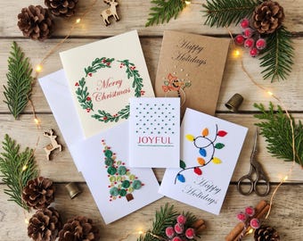 May include: Five greeting cards featuring embroidered holiday designs. One card shows a green wreath with red berries and the text Merry Christmas. Another is a brown card with a snowman and Happy Holidays. A small card says JOYFUL with green dots. A tree card has green circles, and a white card features colorful string lights with Happy Holidays.
