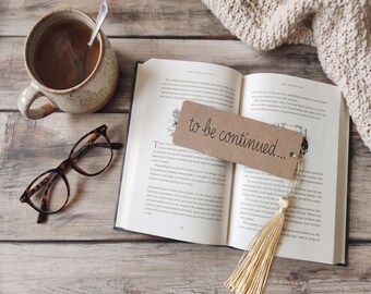 May include: A brown rectangular paper bookmark with the text "to be continued..." printed in black, featuring a gold-colored tassel attached to one end. The bookmark rests on an open book next to a ceramic mug with a spoon and tortoise-shell glasses.